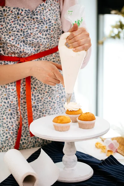 Manos de confitero rematando cupcakes con crema usando una bolsa de pastelería . — Foto de Stock