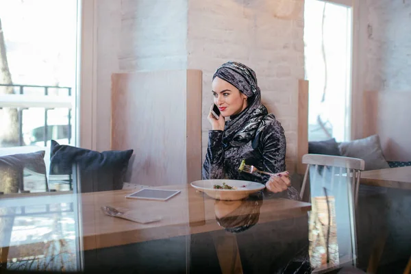 Молодая мусульманка разговаривает по телефону в кафе и смотрит в окно — стоковое фото