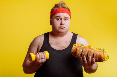 Şişman adam kameraya bakıyor, hamburger ve dambıl tutuyor.