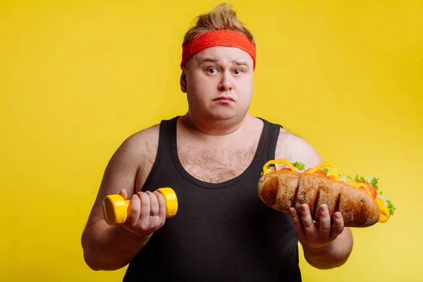 Fat man looking at camera and holding burger and dumbbell — Stock Photo, Image