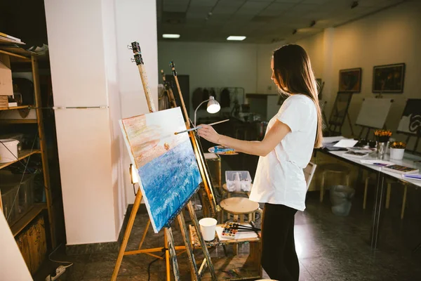 Woman artist painting a picture on easel with oil paints in her workshop