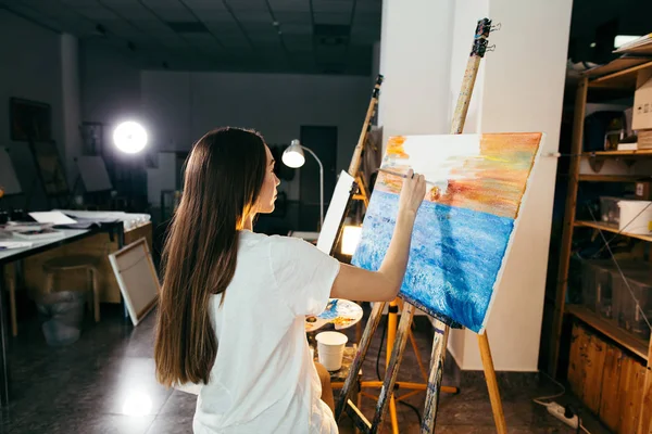 Mujer artista pintando un cuadro sobre caballete con pinturas al óleo en su taller — Foto de Stock