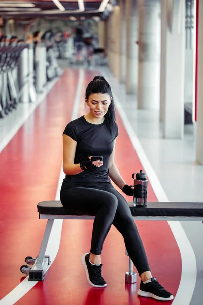 Ajuste mujer con botella usando su teléfono inteligente con auriculares en el gimnasio — Foto de Stock