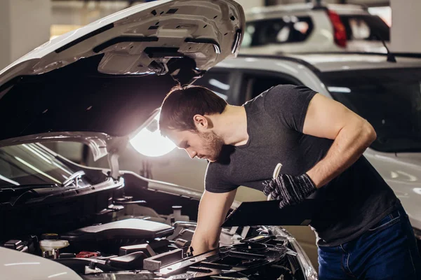 Mechanic examining under hood car and writing notes at garage — Stock Photo, Image