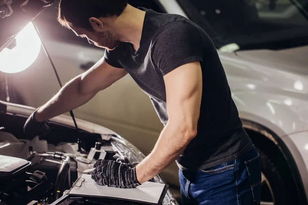 Механик осматривает под капотом машину и пишет заметки в гараже — стоковое фото