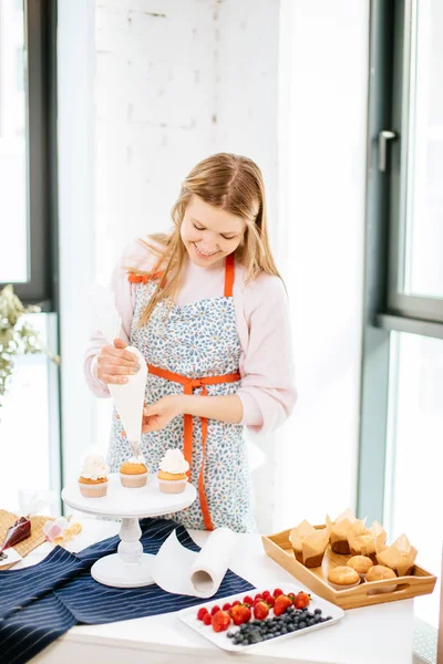 Hermosa mujer pastelera feliz haciendo la parte superior cremosa de cupcakes . —  Fotos de Stock