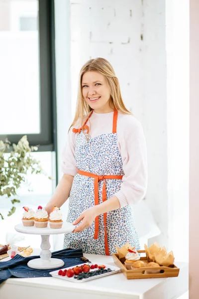 Feliz joven pastelera demostrando cupcakes recién horneados mirando a la cámara y sonriendo —  Fotos de Stock