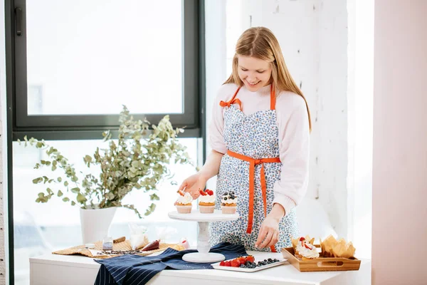 Jeune femme au foyer blonde décorer des cupcakes avec des baies sur le plateau de service en cuisine — Photo