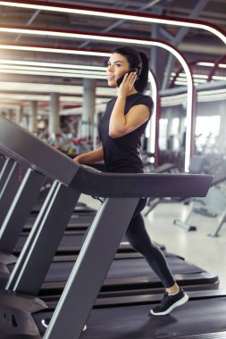 Kadın koşu bandında koşarken cep telefonuyla konuşurken gülümsüyor.
