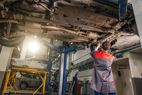 Mecánico debajo del coche en garaje examinando neumático y condición técnica — Foto de Stock