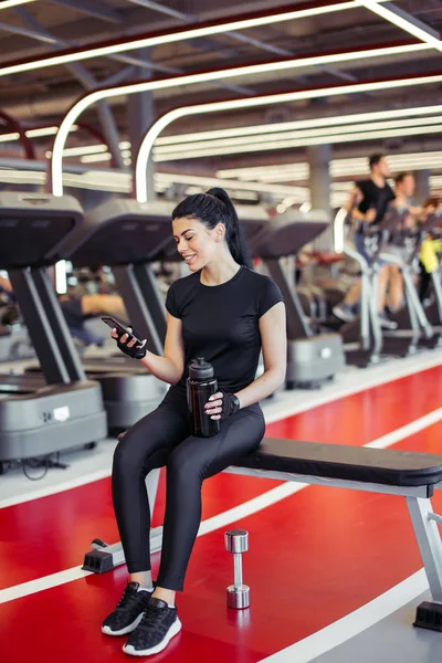 Fit wanita dengan botol menggunakan ponsel pintar dengan earphone di gym — Stok Foto