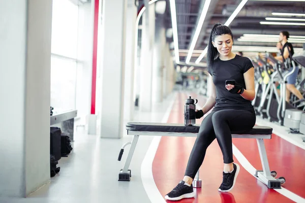 Fit vrouw met fles met behulp van haar smartphone met oortjes in de sportschool — Stockfoto