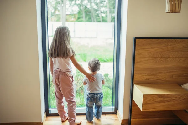 Küçük kız ve erkek kardeş. Çocuklar arkada, panoramik pencerenin önünde duruyorlar. — Stok fotoğraf