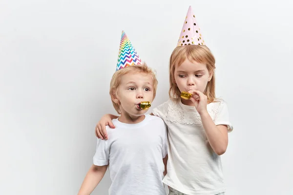 Двое милых детей используют свистки для вечеринок, носят шляпы на белом фоне — стоковое фото