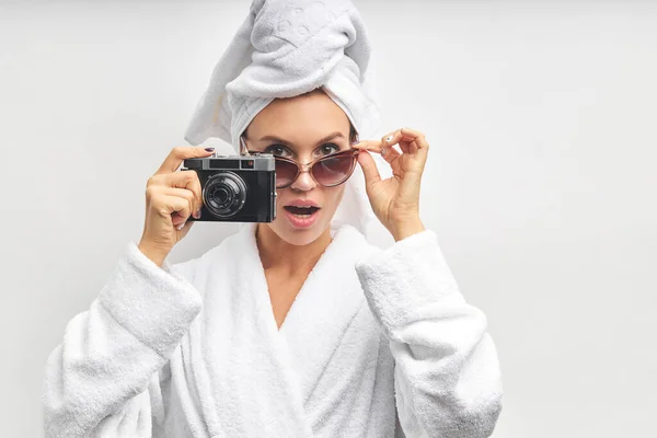 Aufgeregtes und überraschtes Mädchen im Bademantel steht isoliert vor weißem Hintergrund und hält Fotokamera — Stockfoto