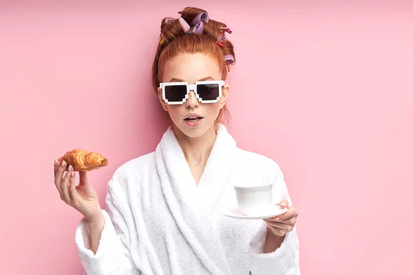 Linda chica pelirroja juguetona en gafas de sol, con albornoz y beber té con croissant — Foto de Stock