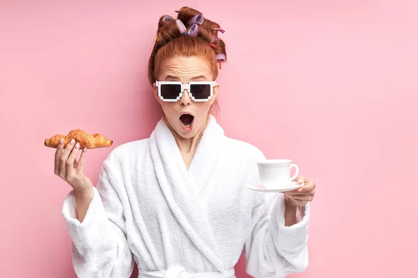 Sorprendido joven pelirroja en bata de baño de pie aislado sobre fondo rosa, la celebración de croissant — Foto de Stock