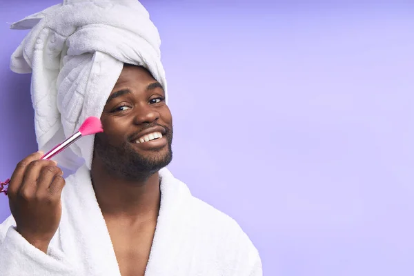 Hombre africano divertido usando albornoz con cepillo de maquillaje mirando a la cámara — Foto de Stock