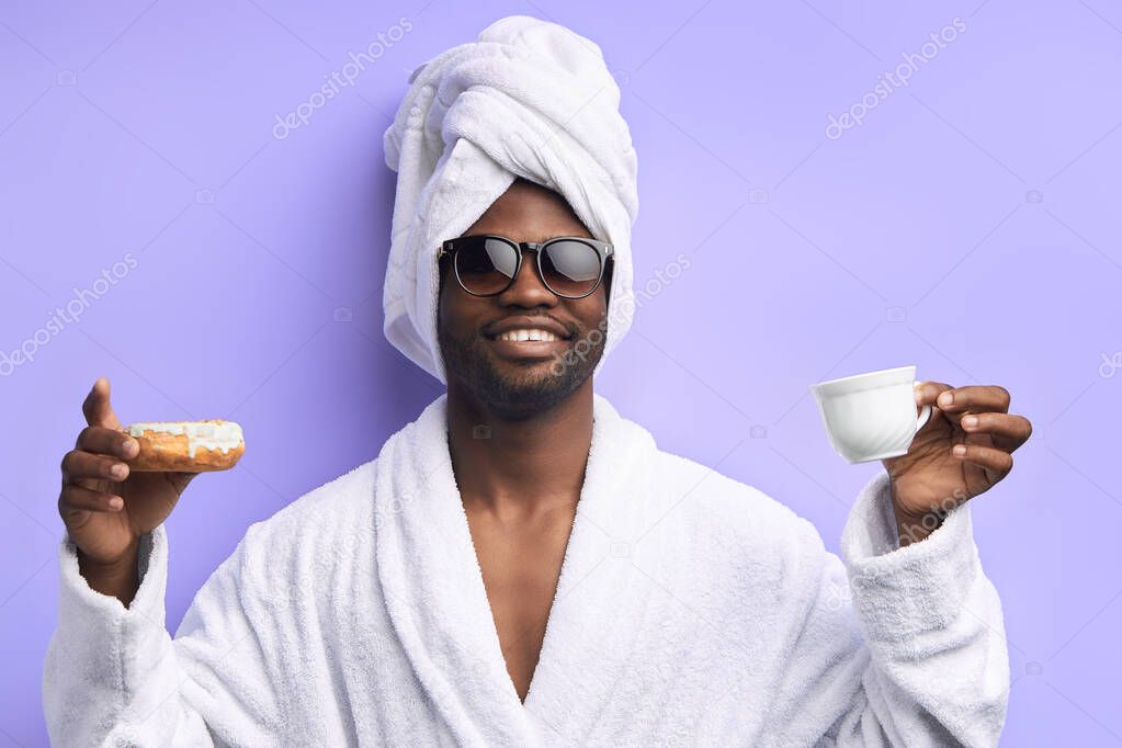 Excited young african man holding glazed donut and cup of tea or coffee