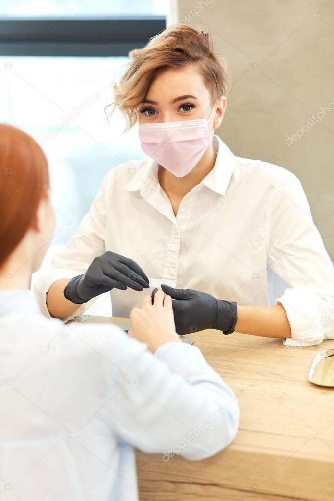 Masked master manicurist looking at camera while working with clients nails at beauty salon