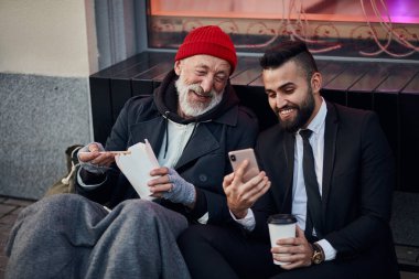 Young kind-hearted businessman and senior beggar look at phone clipart