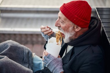 Old-aged vagabond sitting on walking street and hungrily eating clipart
