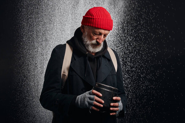 Bum with grey beard stand under heavy rain with cup for money