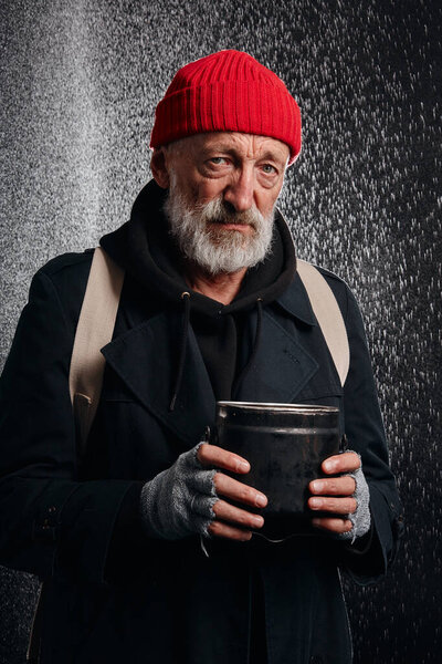 Склоняющийся старый нищий в уличной одежде стоит под ливнем
