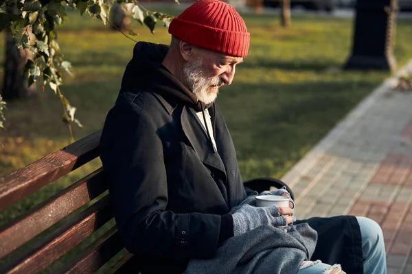 Persona anziana senza casa, soldi e lavoro seduta su panchina in strada con tazza per soldi — Foto Stock