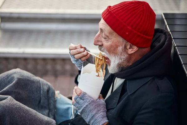 Старый бродяга сидит на пешеходной улице и голодно ест — стоковое фото