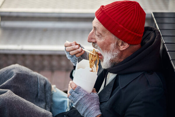 Old-aged vagabond sitting on walking street and hungrily eating