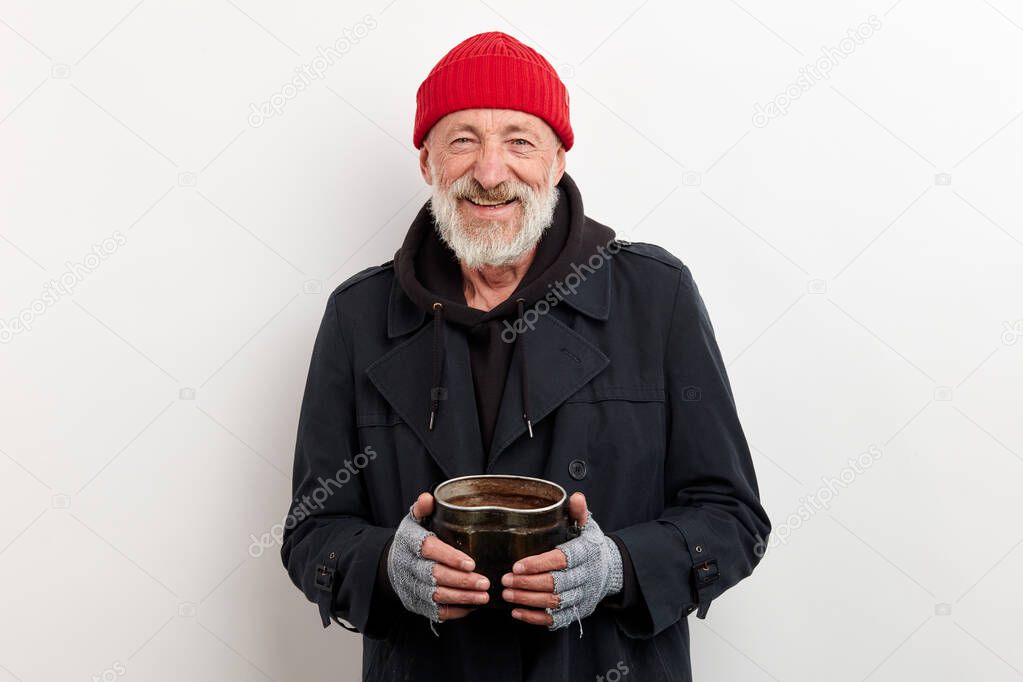 Positive old homeless man with grey beard in black coat holding iron can for money