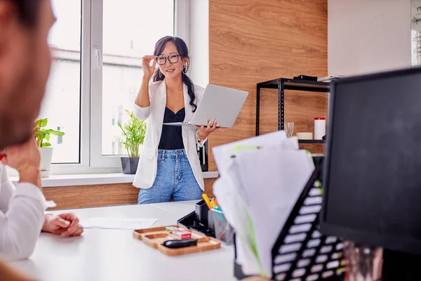 Knappe aziatische vrouw in bril houden laptop terwijl praten voor groep van mensen — Stockfoto