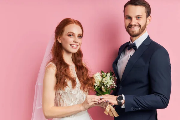 Homem caucasiano com barba em smoking com arco colocando anel no dedo de sua namorada em vestido de noiva — Fotografia de Stock