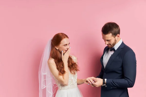Mulher feliz e homem vestindo smoking e vestido de casamento branco isolado sobre o chão rosa — Fotografia de Stock