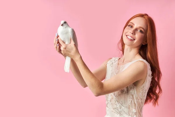 Young female with red hair in white dress holding white dove — Stock Photo, Image
