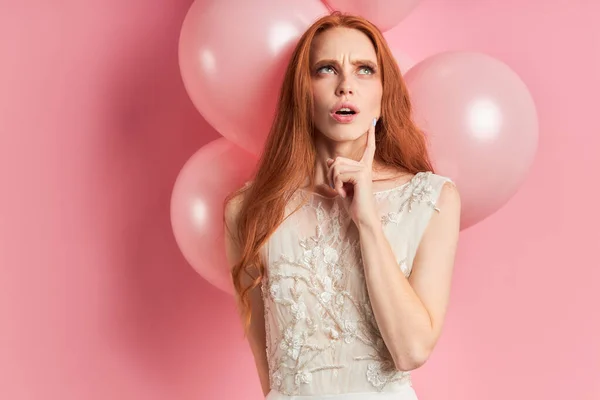 Thoughtful redhead woman in wedding dress isolated over pink background