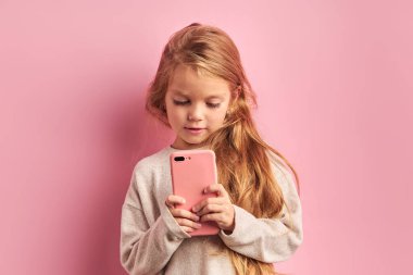Pembe arka planda, cep telefonunu elinde tutan sevimli küçük kız.