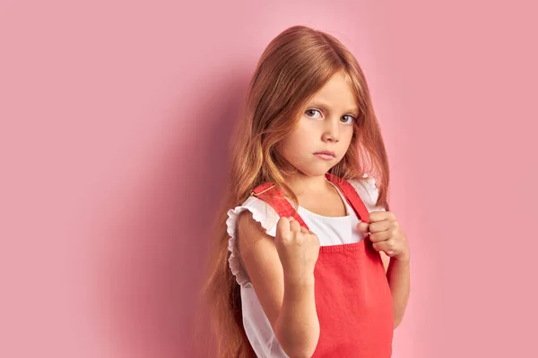 Szomorú kislány piros overallban elszigetelt rózsaszín háttér — Stock Fotó
