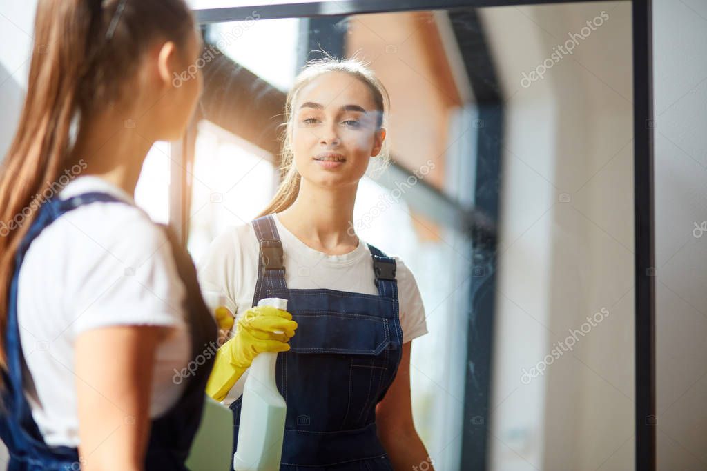 Young woman as professional cleaner in uniform cleaning mirror