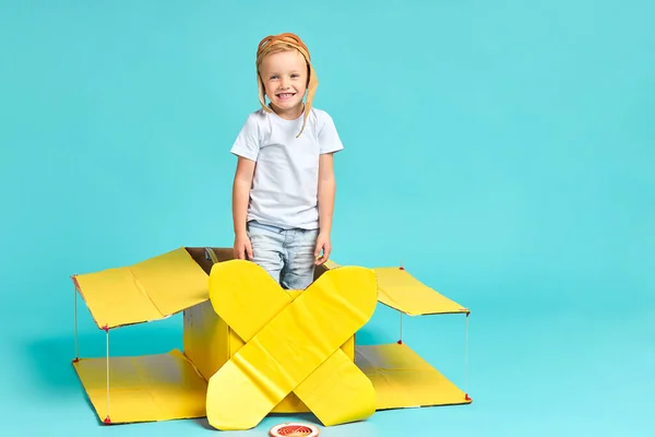 Мальчик играет с желтым картонным самолетом, изолированным на голубом — стоковое фото