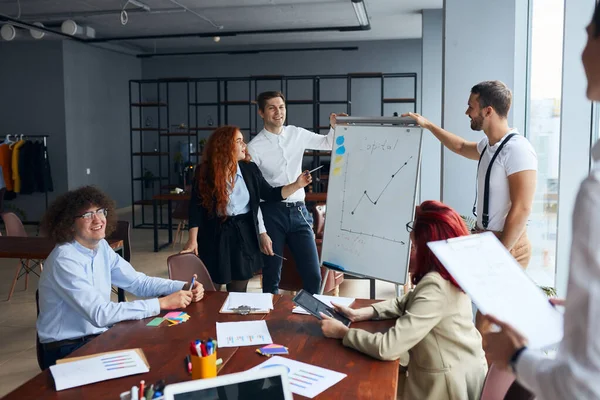 Giovane manager che spiega il progetto di business su flip chart al collega in ufficio moderno — Foto Stock