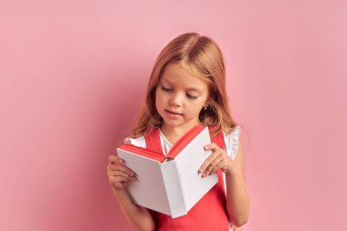Akıllı kız, pembe arka planda izole edilmiş kitabı dikkatle okuyor.