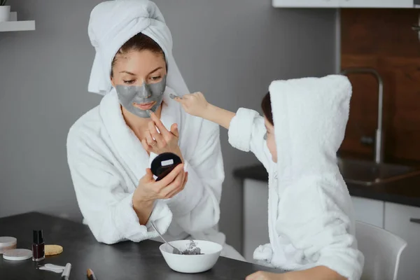 Bewonderenswaardige vrouw en kind in handdoeken en badjassen gebruik schoonheid maskers — Stockfoto