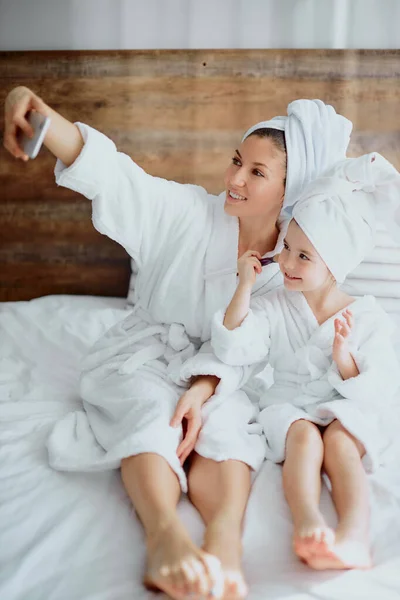 Hermosa mujer y niño pequeño tomando selfie usando albornoz en la cama — Foto de Stock