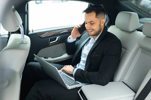 Junger Geschäftsmann arbeitet am Laptop und spricht im Auto mit dem Smartphone — Stockfoto