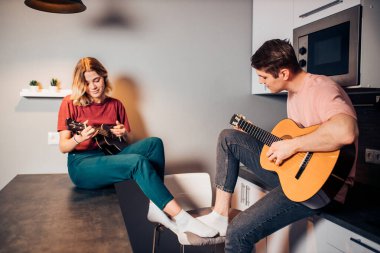 Beyaz çocuk gitar çalıyor ve kadın mutfakta ukulele çalıyor.