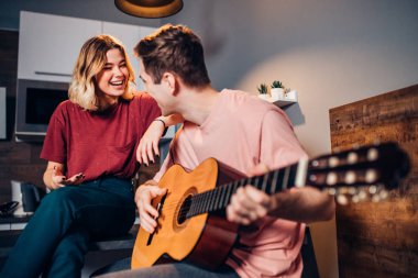 İki beyaz insan evde birlikte gitar çalıyor.