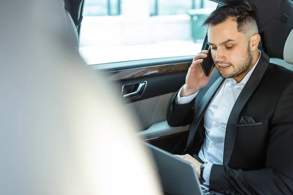 Junger Geschäftsmann arbeitet am Laptop und spricht im Auto mit dem Smartphone — Stockfoto