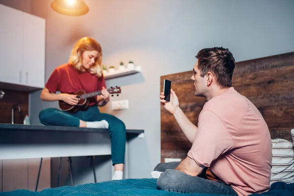 beautiful woman playing ukulele at home while his boyfriend take photo or shoot video